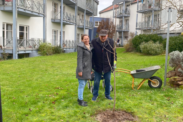 Dudenhofen: Stellvertretende Pflegedienstleiterin neben Bewohner und Kirschbaumpate vor Baum.